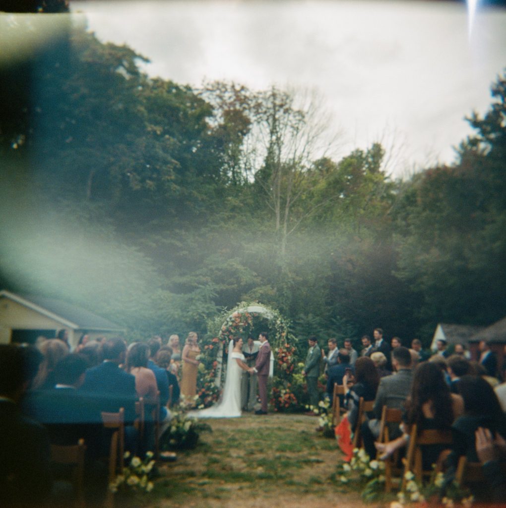 Film photograph of backyard wedding ceremony; bride and groom stand with their hands clasped while wedding guests look on