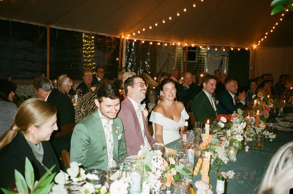 Bride and groom sit with their guests at reception in backyard tent with lush floral decorations