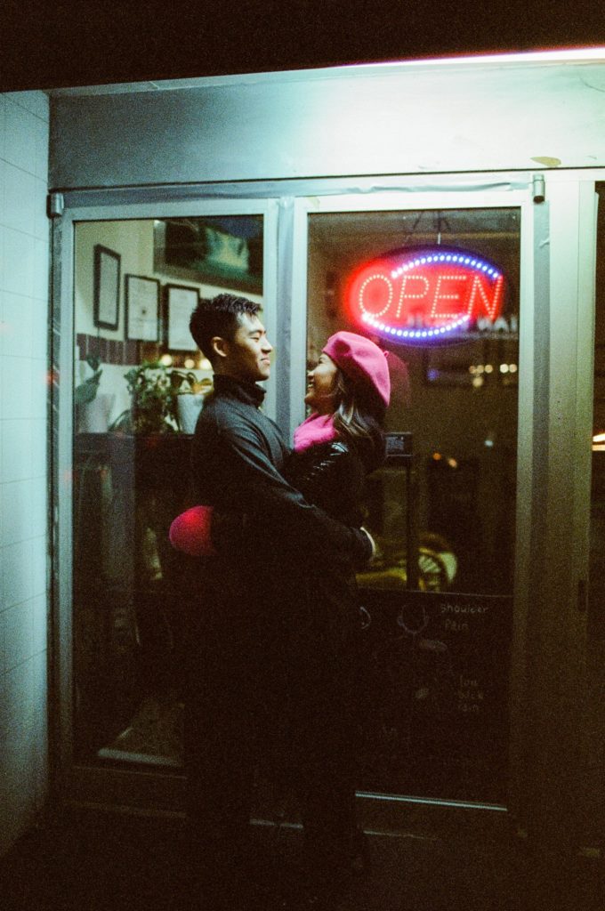 Film photo of a couple smiling and hugging in front of a storefront window at night in Brooklyn