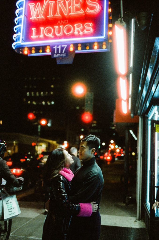 New York neon lights illuminate an engaged couple in each others' arms on the street