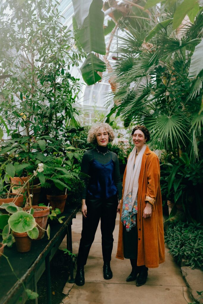 Two women pose and smile together celebrating their engagement in a greenhouse
