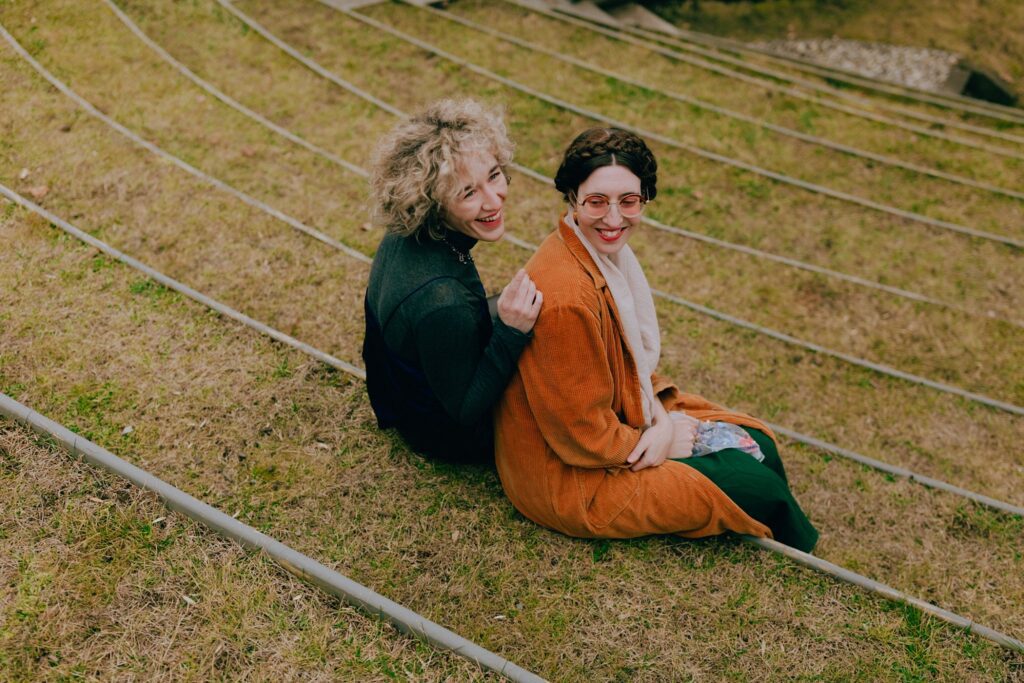 An engaged couple sits on grass and smiles back at the camera