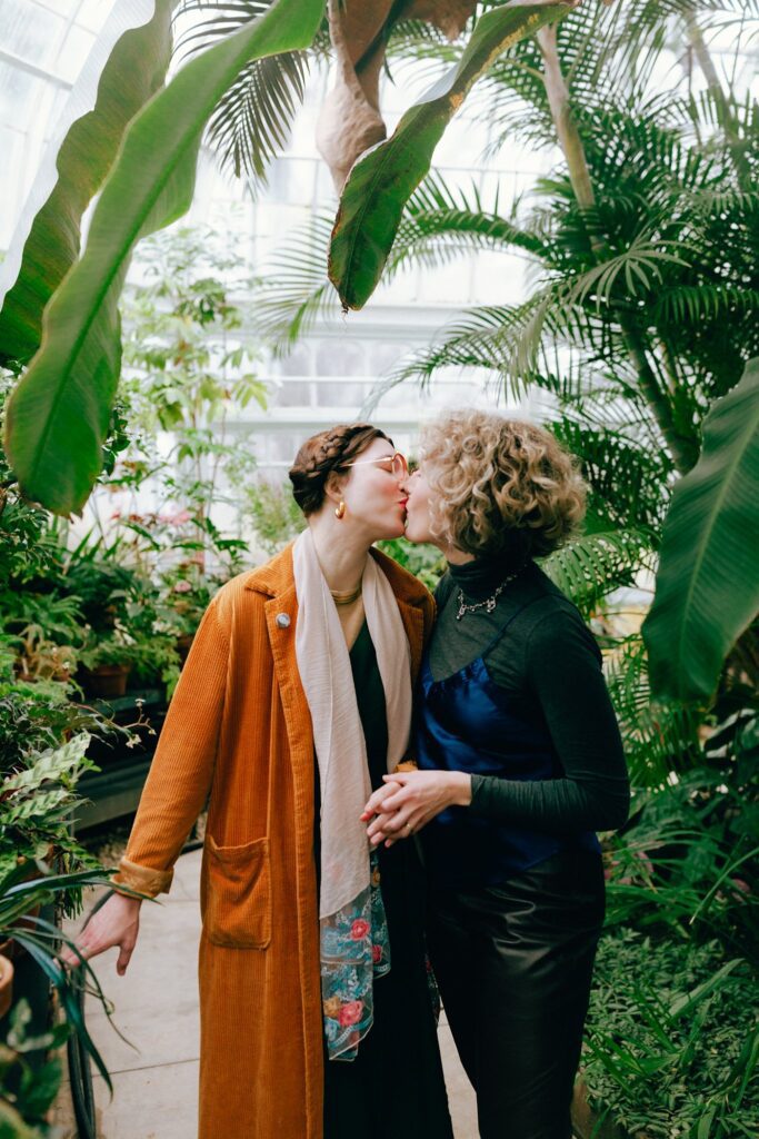 Two women kiss and hold hands among various plants