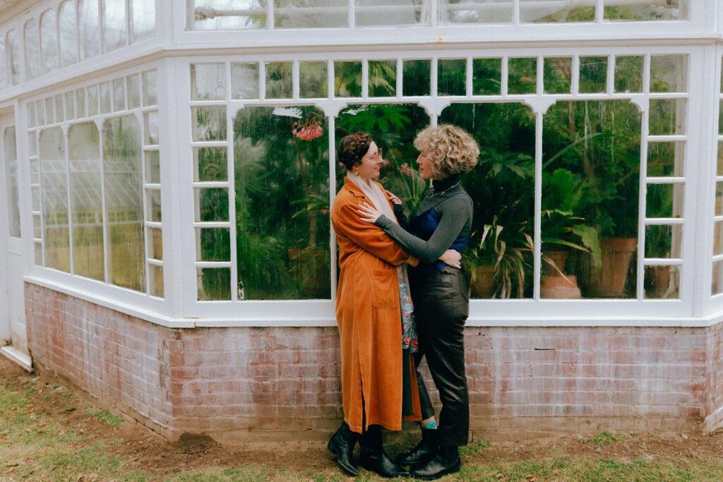 Same sex couple holds each other in front of a greenhouse window