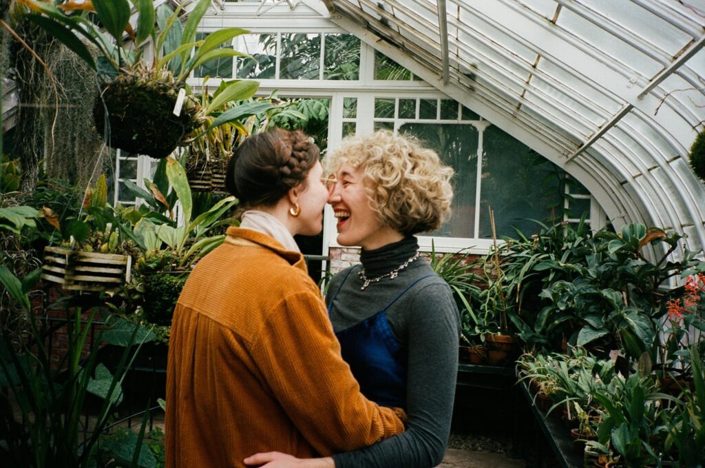 Same sex couple laughs together while embracing in a greenhouse