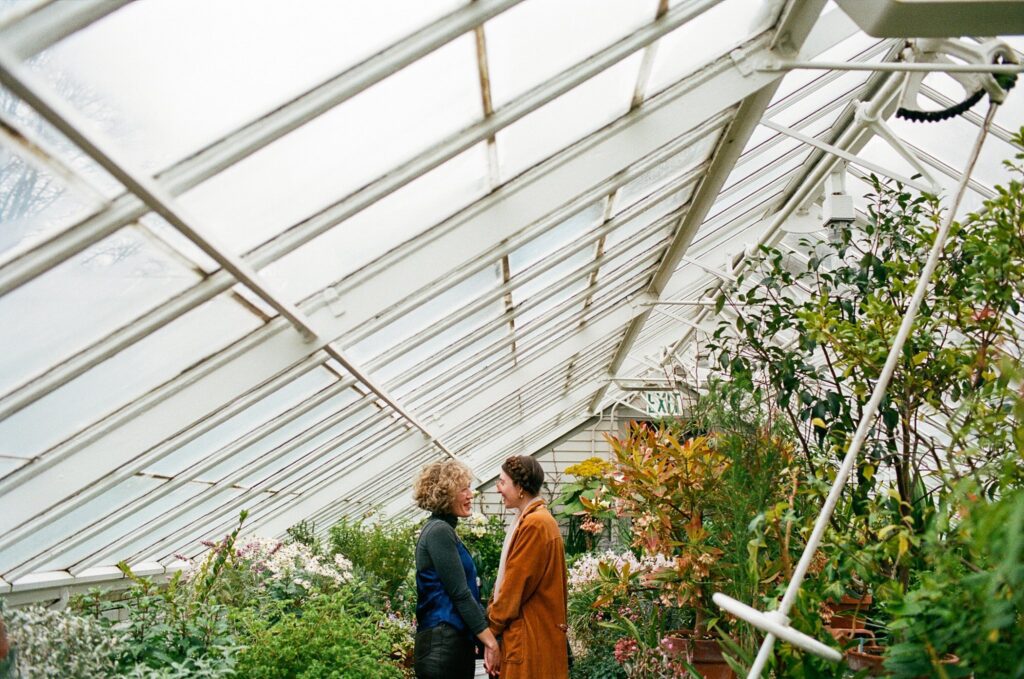 A wide variety of plant life grows all around a happy couple while light pours in through the greenhouse roof