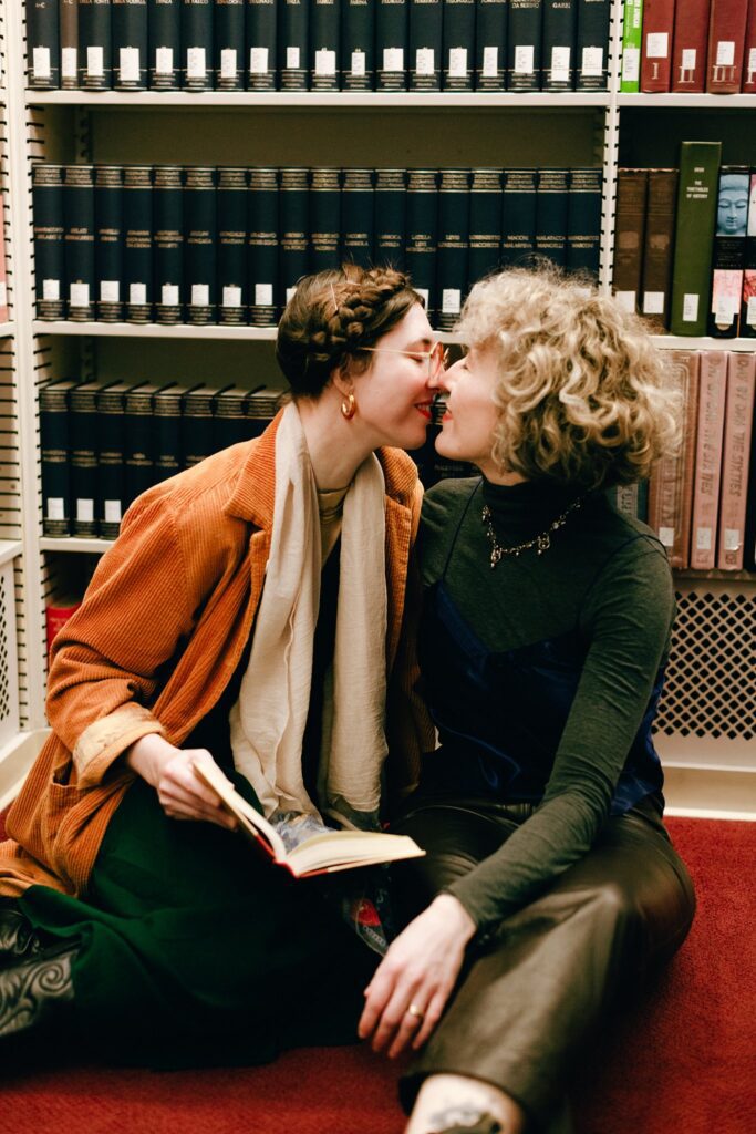 Seated on the floor of a library in front of shelves full of books, a woman leans in to kiss her future bride while holding an open book.