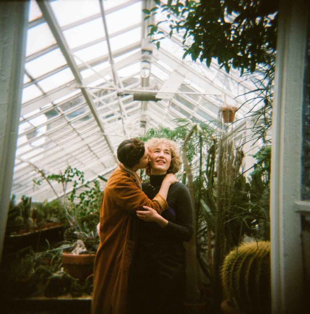 Fiancée kisses her bride-to-be on the cheek inside a greenhouse, captured on film