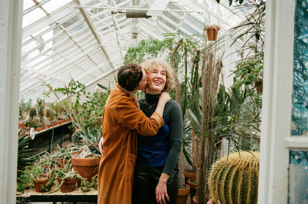 Woman kisses her bride-to-be on the cheek in front of greenhouse plants