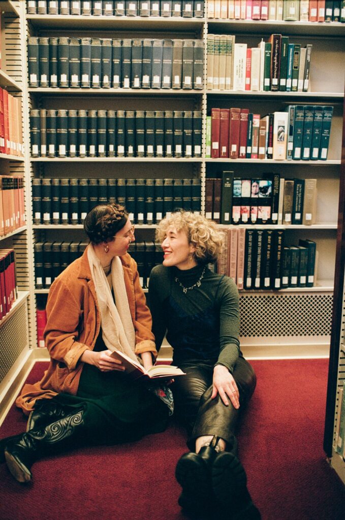 Two women sit together and smile in front of a bookshelf
