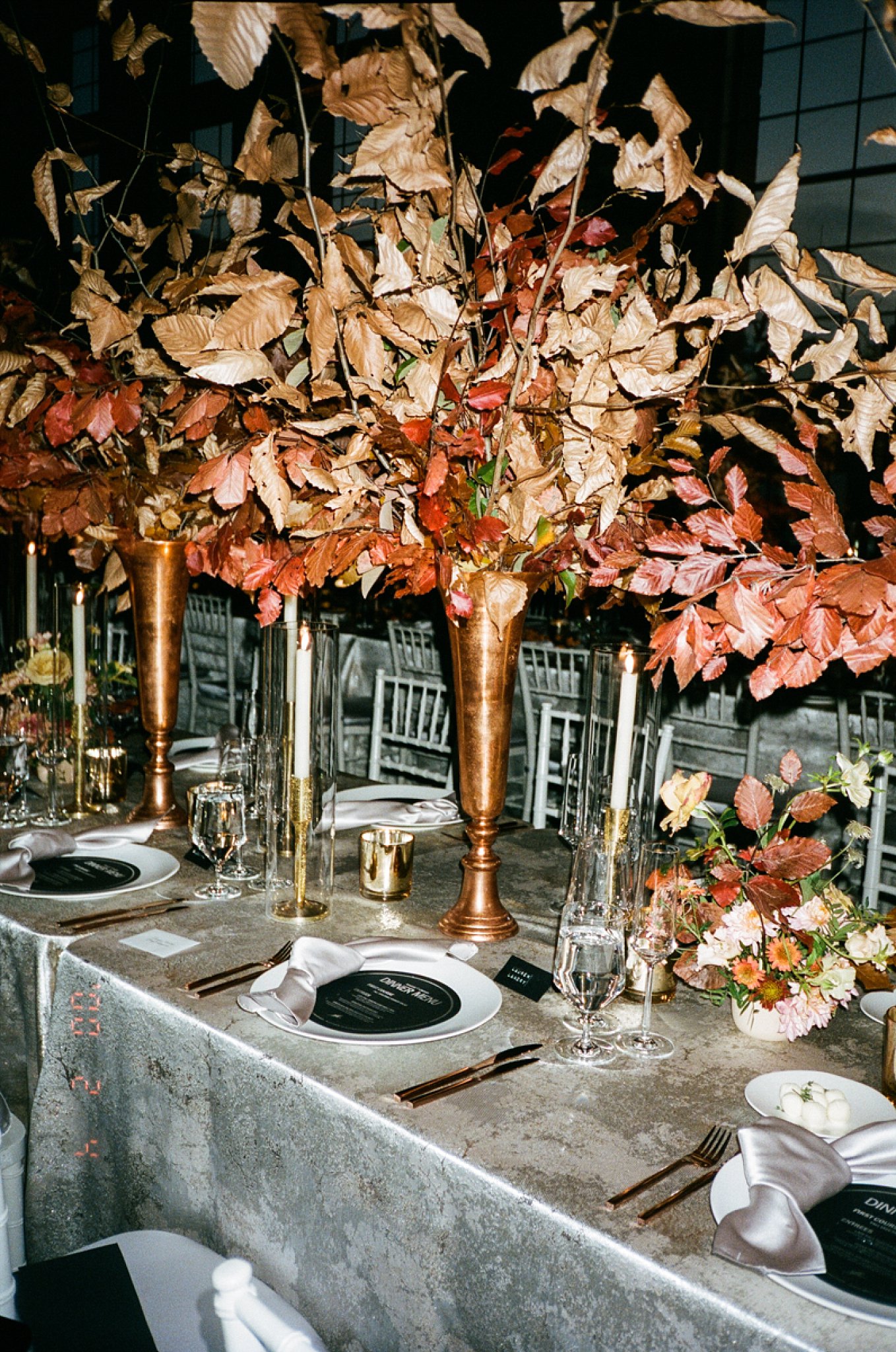 Details of a tablescape at an artistic Boston Wedding, long dinner table with place settings, flutes, glasses, candles and tall brass vases with leaves.