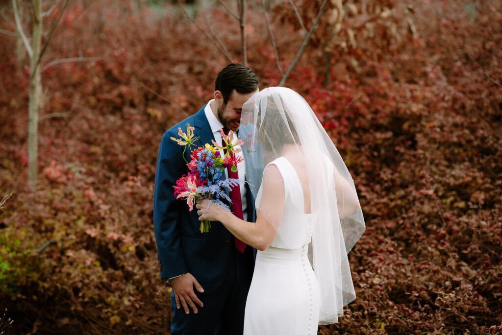 Groom smiles as his bride moves close to him, set against Massachusetts fall foliage