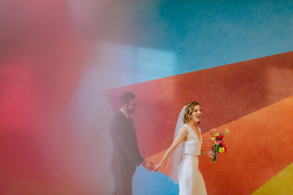 Bride and groom laugh and walk in front of brightly colored piece of art