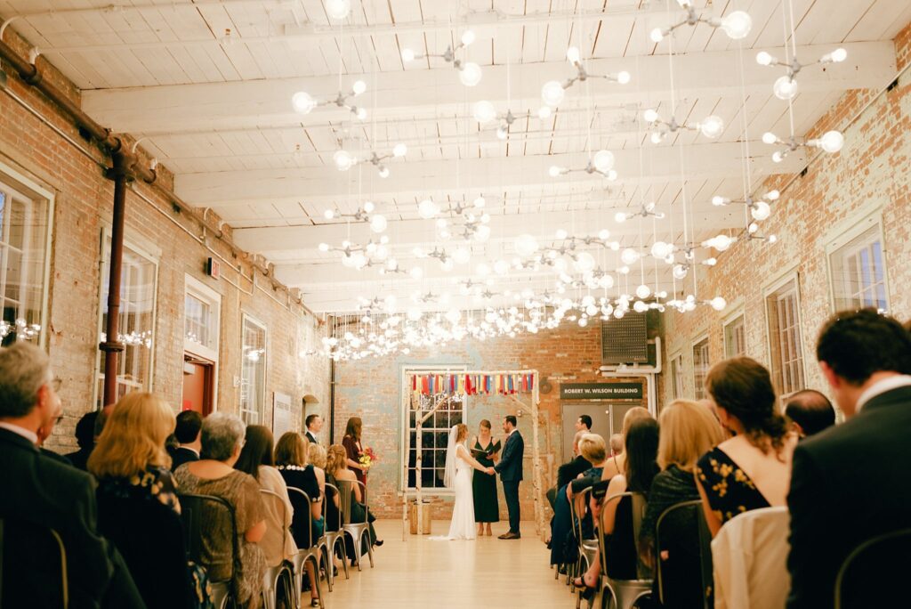 Lights glow overhead in Mass MoCA during wedding ceremony while bride and groom hold hands in front of all their guests