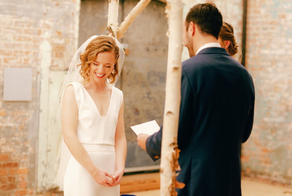 Bride smiles at her wedding ceremony while groom reads his vows