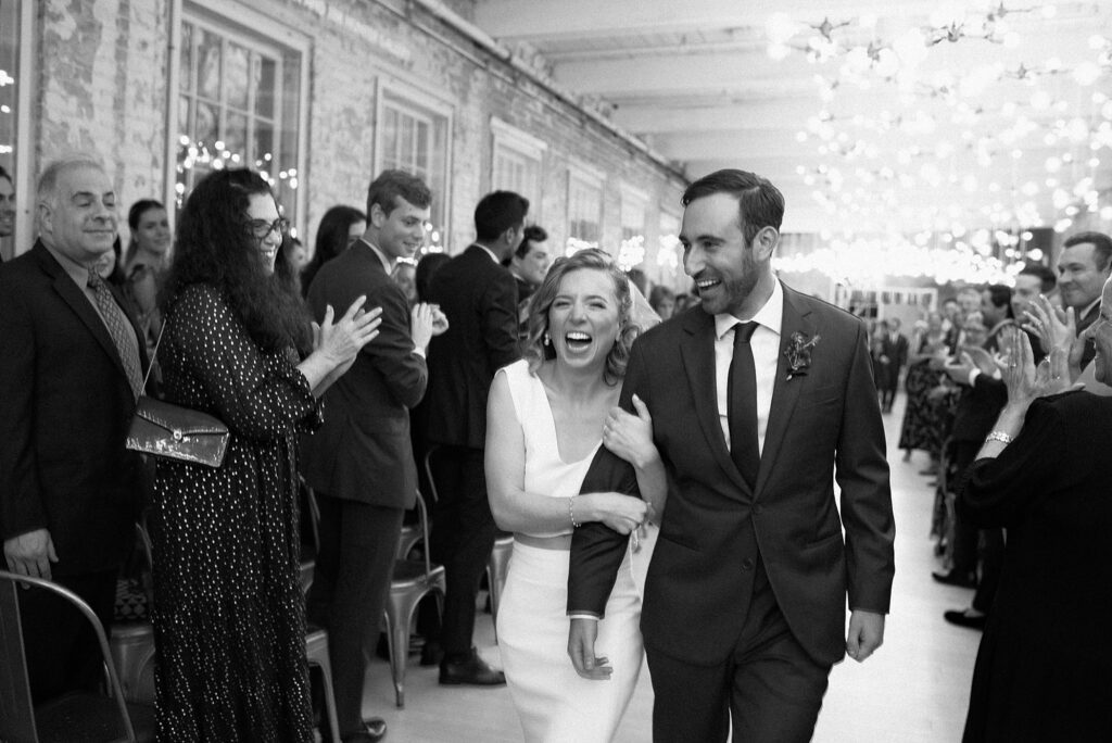 Bride laughs while guests clap immediately following wedding ceremony, in black and white