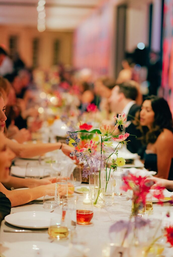 Vibrant colors adorn a photo of table flowers with seated guests conversing in the background