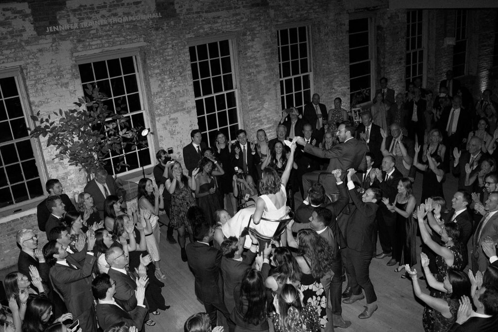Black and white overhead shot of newlyweds being hoisted on chairs above the dance floor by guests