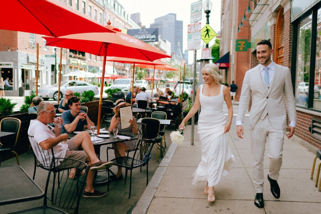 a couple walks by people eating at a restaurant on their wedding day