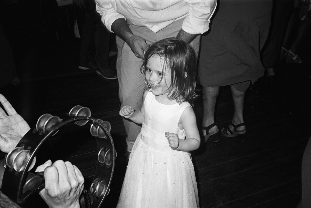 Joyful black and white film photograph of a young girl smiling on the dance floor, a tambourine playfully visible in the foreground, capturing the energy of the wedding celebration.