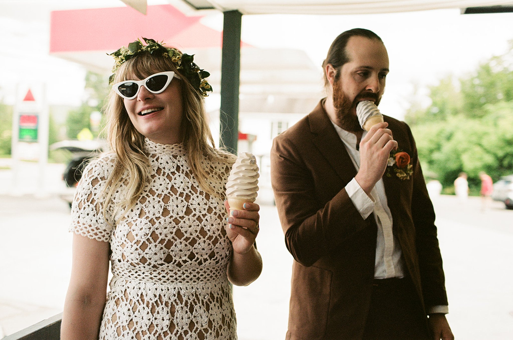 Bride and groom enjoy soft serve ice cream in wafer cones together, she smiles while he goes in for a bite.