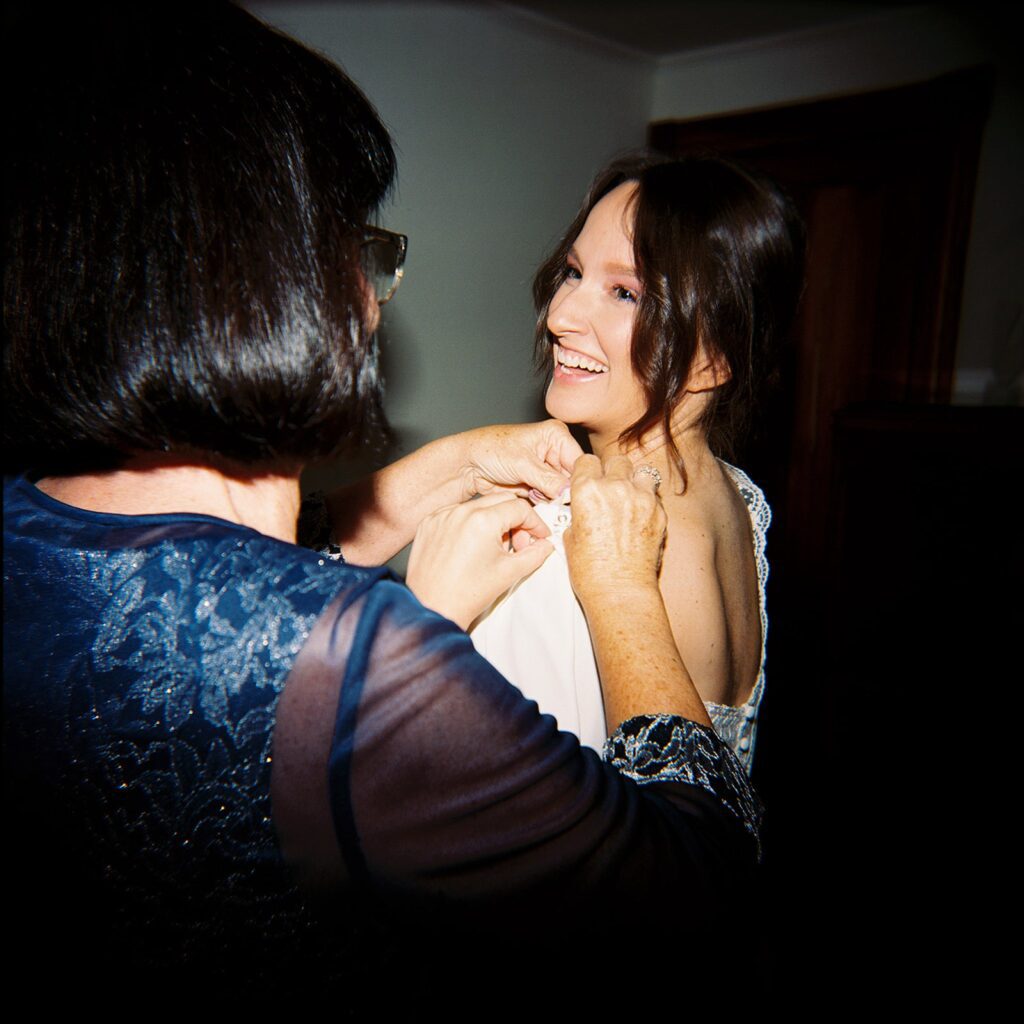 a bride laughs over her shoulder as her wedding dress is being put on