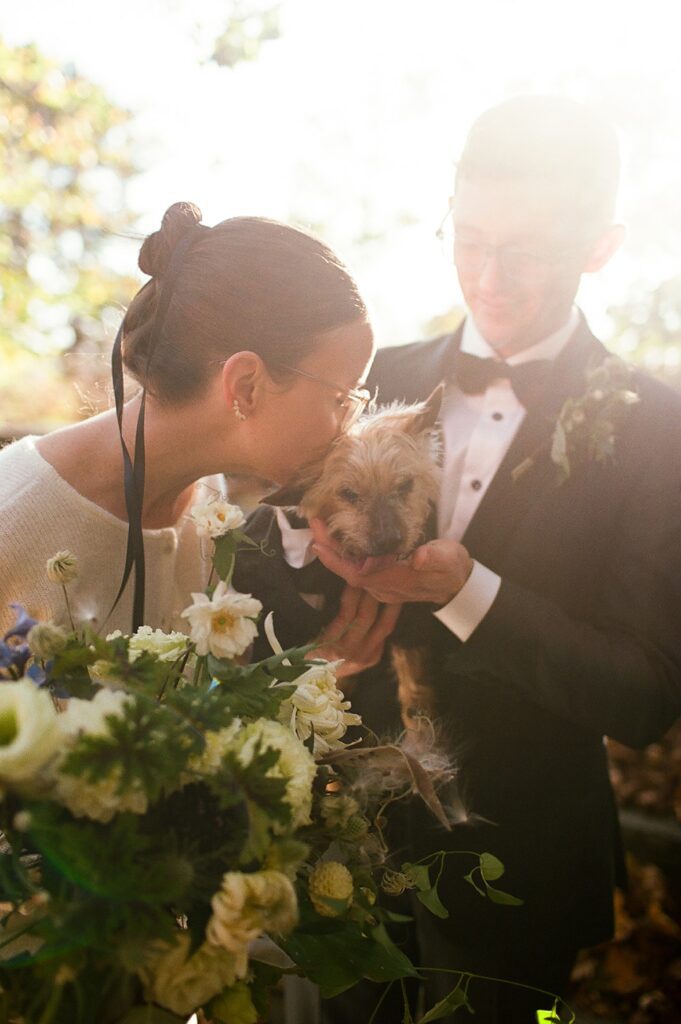 Bride kisses her dog while groom holds in while sun shines from above, shot on film